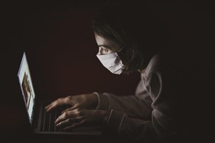 A person wearing a face mask looks at their laptop in the dark